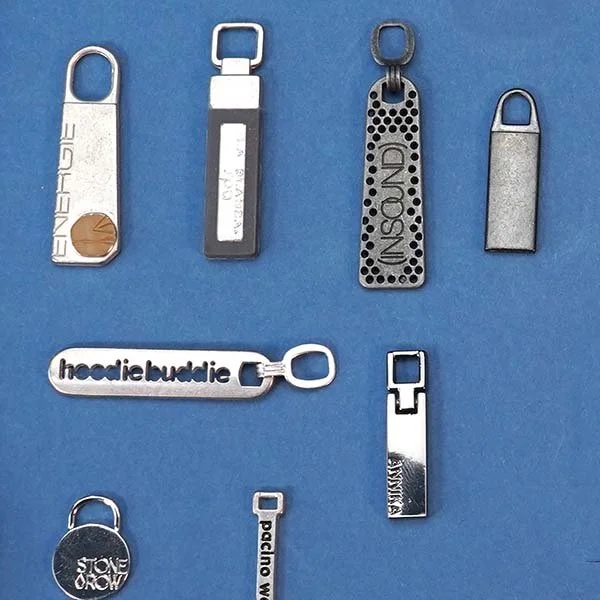 Eight metal zipper pulls with various designs and text, laid out on a blue background.