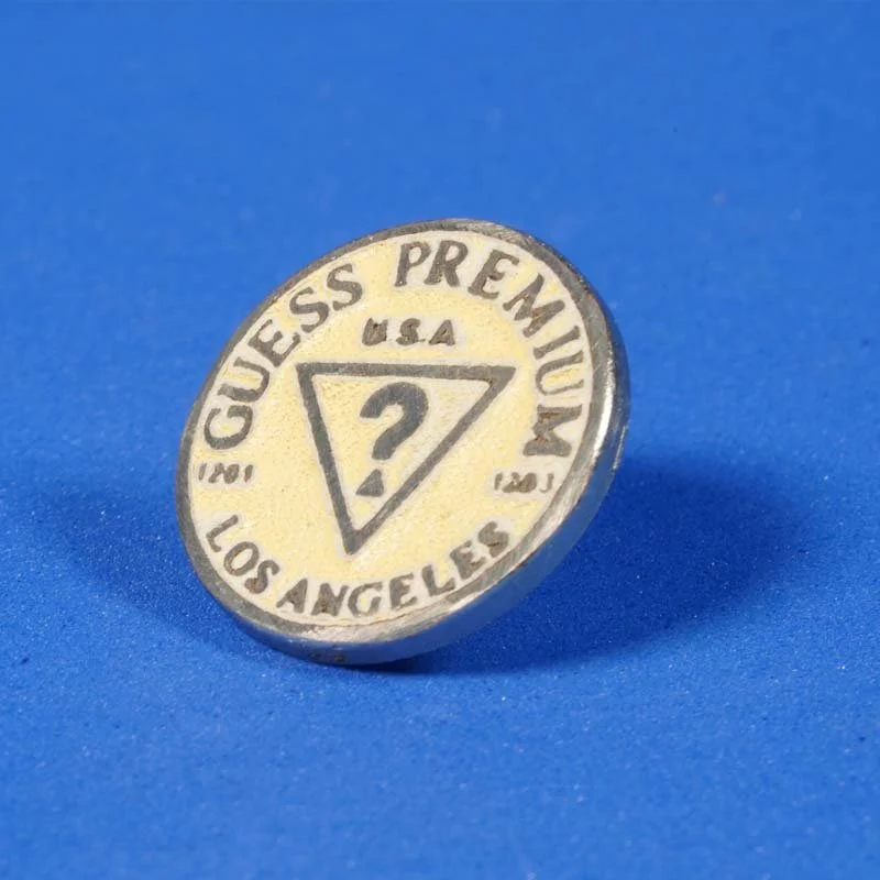 Close-up of a vintage Guess Premium button badge with a yellow background, black text, and a question mark inside a triangle, against a blue background.