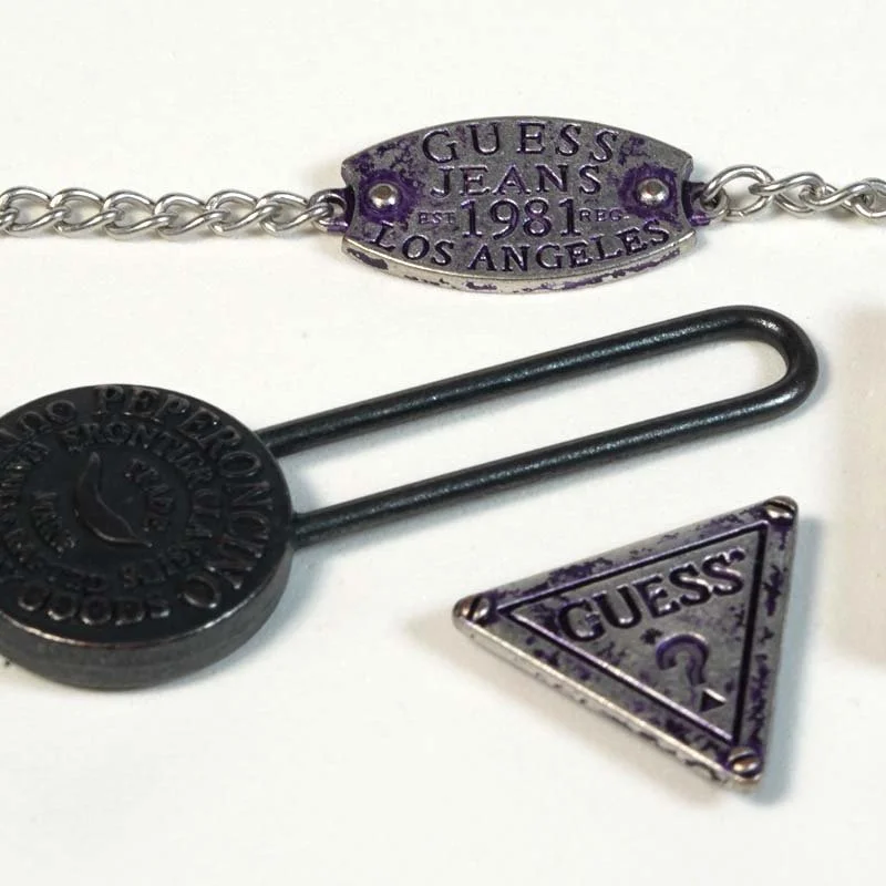 Close-up of three Guess accessories: a bracelet with a purple metal plate, a black circular pin with Guess logo, and a triangular pin with Guess logo and question mark.