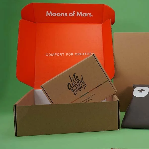 Open box from Moons of Mars with a smaller cardboard box inside labeled 'Life Around Angels' and a gray object with a plus sign on it next to the box, all on a green background.