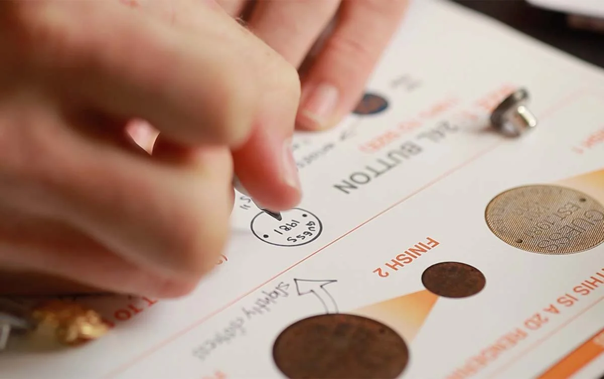 A person is marking a ballot paper with a black pen for an election, with several other marked and unmarked coins and a metallic object nearby.