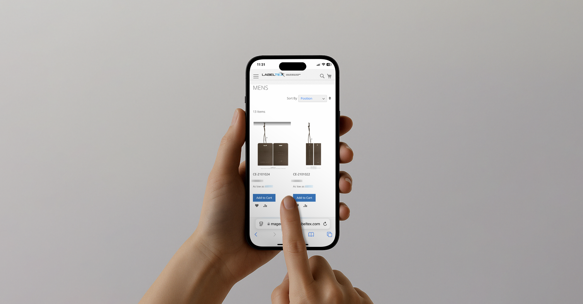 Close-up of a person's hand holding a smartphone displaying an online shopping website with men's products, including two black tags with item numbers and 'Add to Cart' buttons.