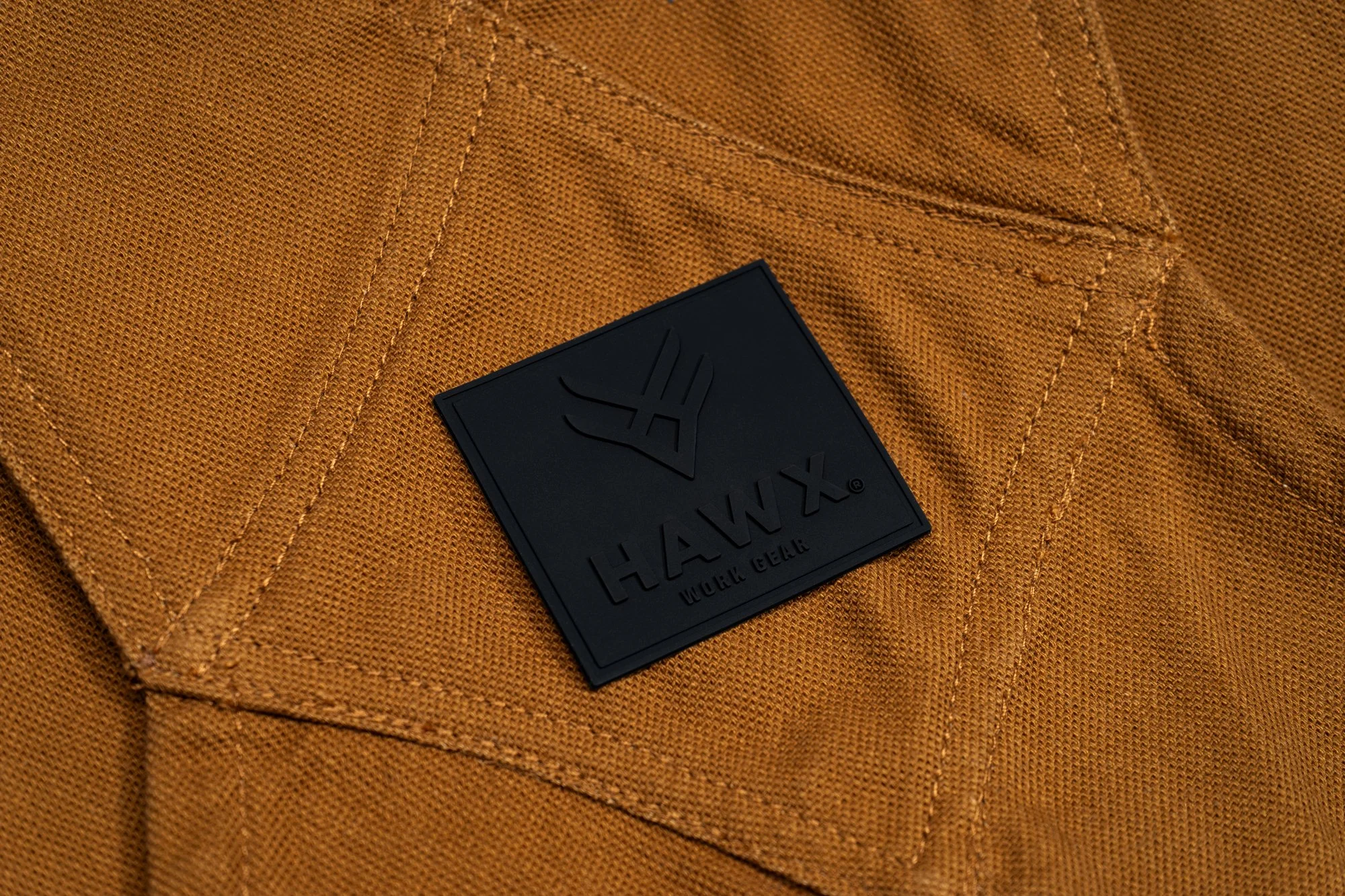 Close-up of an orange work jacket with a black square Hawx logo patch sewn onto the chest pocket.