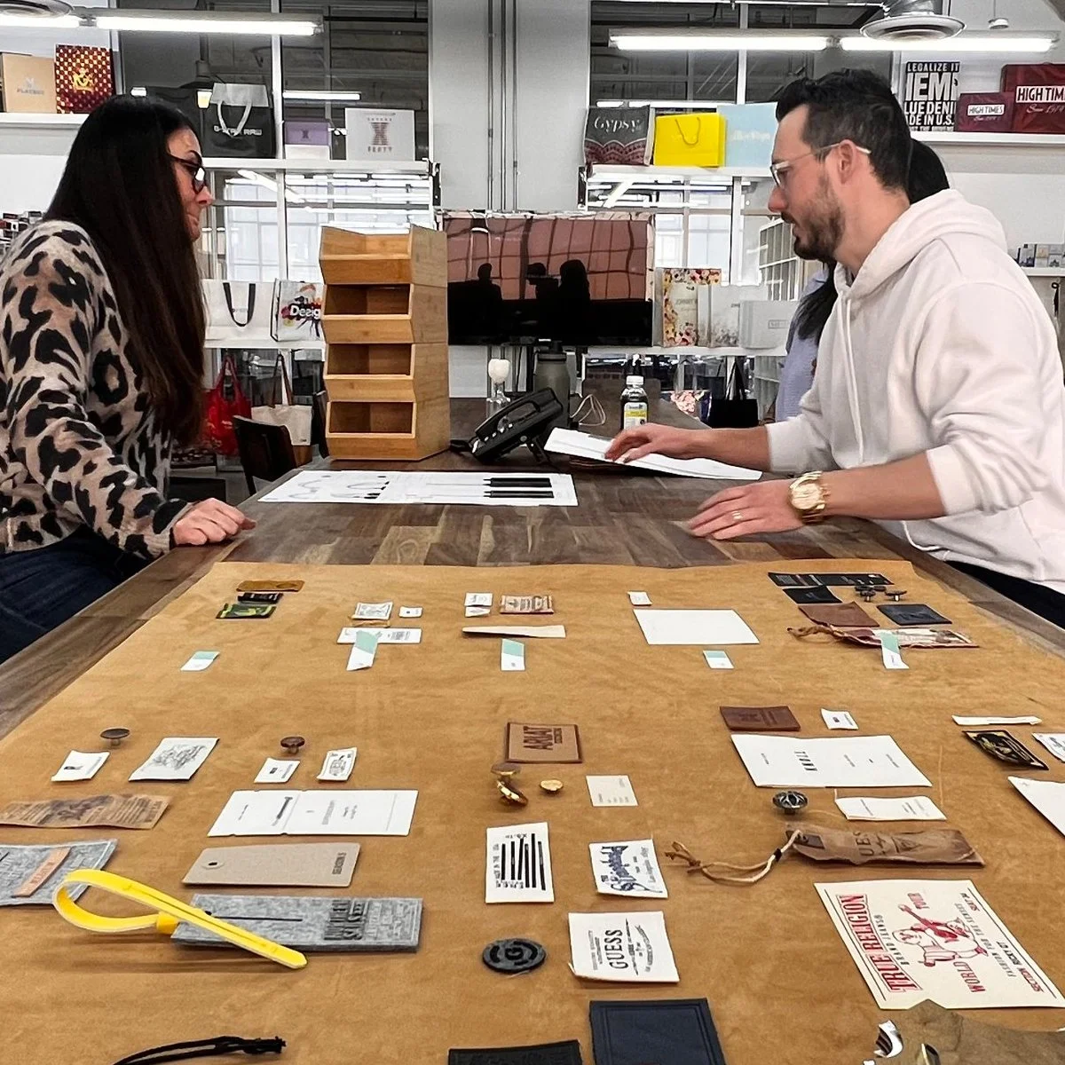 Two people playing a board game at a table in a retail or hobby store. The table has various game pieces, cards, and tokens. One person, wearing a leopard print sweater, is facing another person in a white hoodie, who is holding a book or game manual. There are shelves and boxes in the background.