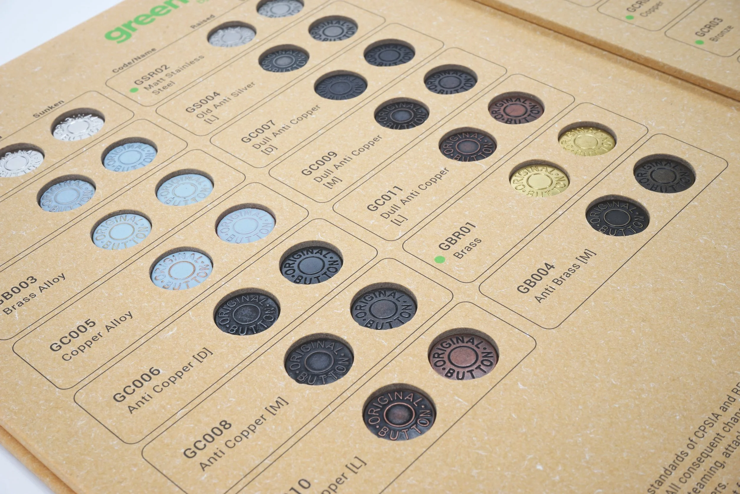 A display sheet of various colored metal buttons, labeled with descriptions such as 'GSR02 Matt Stainless Steel,' 'GSS04 Old Silver,' and 'GC001 Dull Anti Copper,' arranged in rows and columns.