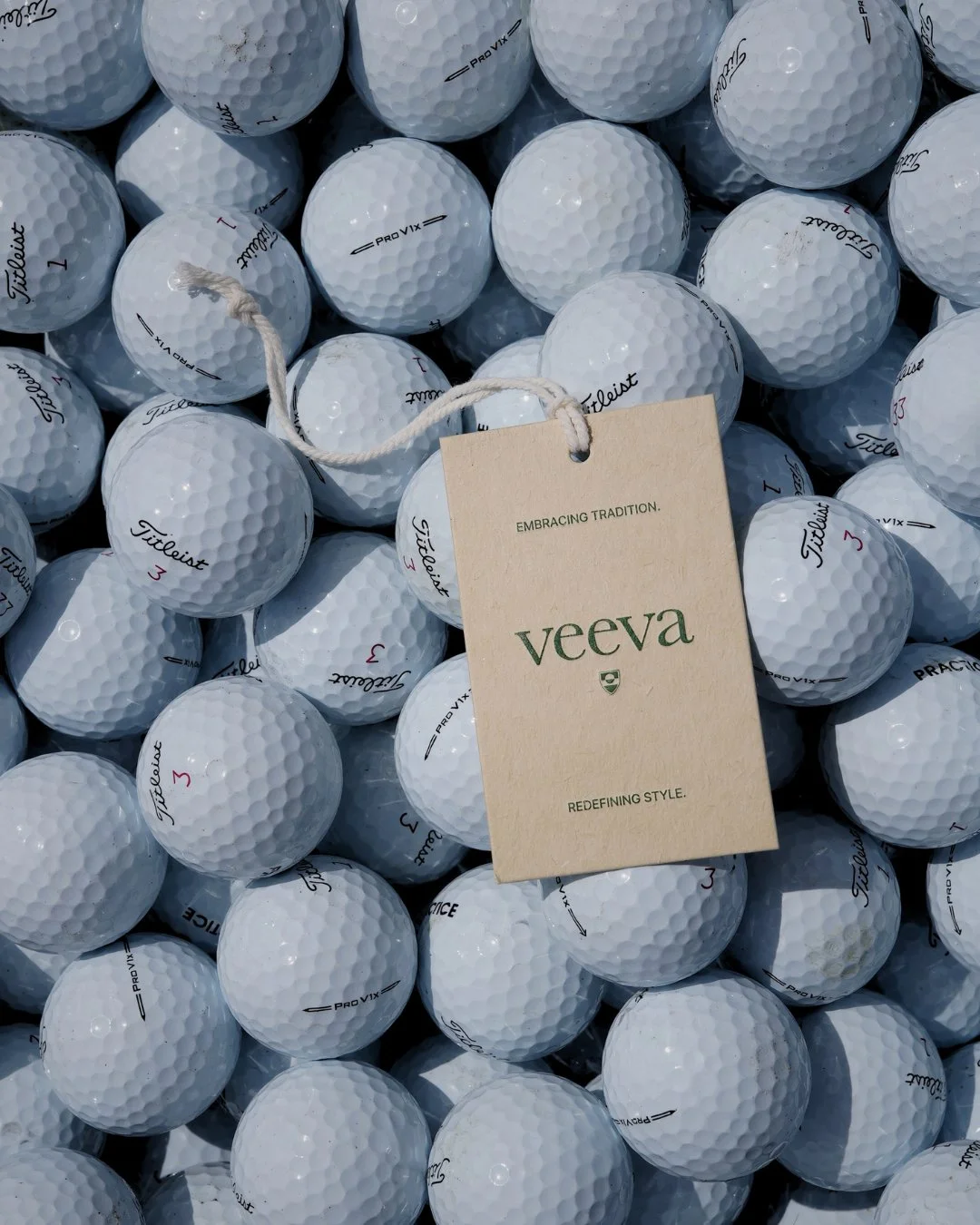 Group of white Titleist golf balls with black and red markings, some with a tag reading 'Veeva,' and a beige card with the text 'Embracing Tradition. Veeva. Redefining Style.'