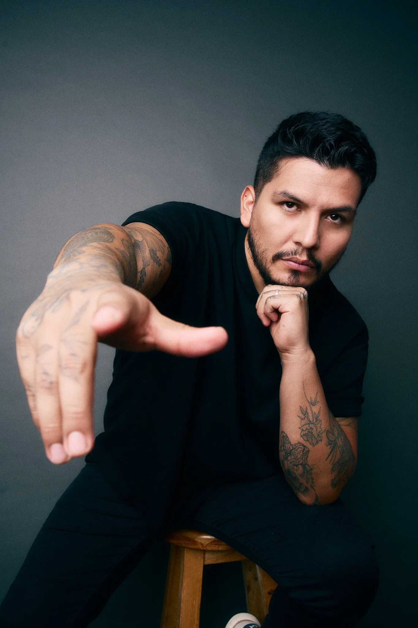 A man with tattoos and dark hair wearing a black t-shirt sitting on a stool, reaching toward the camera with a serious expression, against a dark gray background.