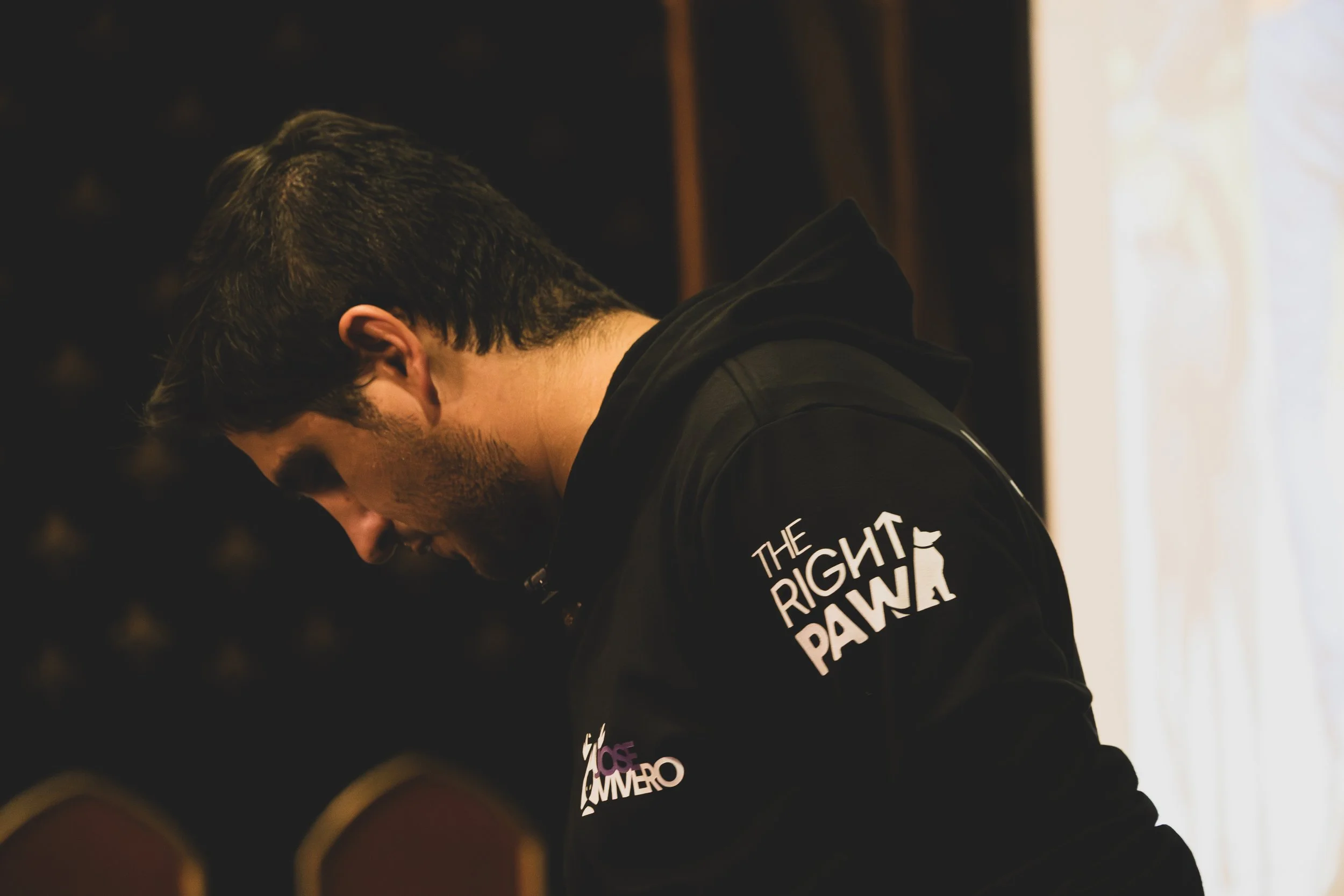 Man with dark hair and glasses wearing a black jacket with the logo 'The Right Paw' on the sleeve, looking downward in a dimly lit room.