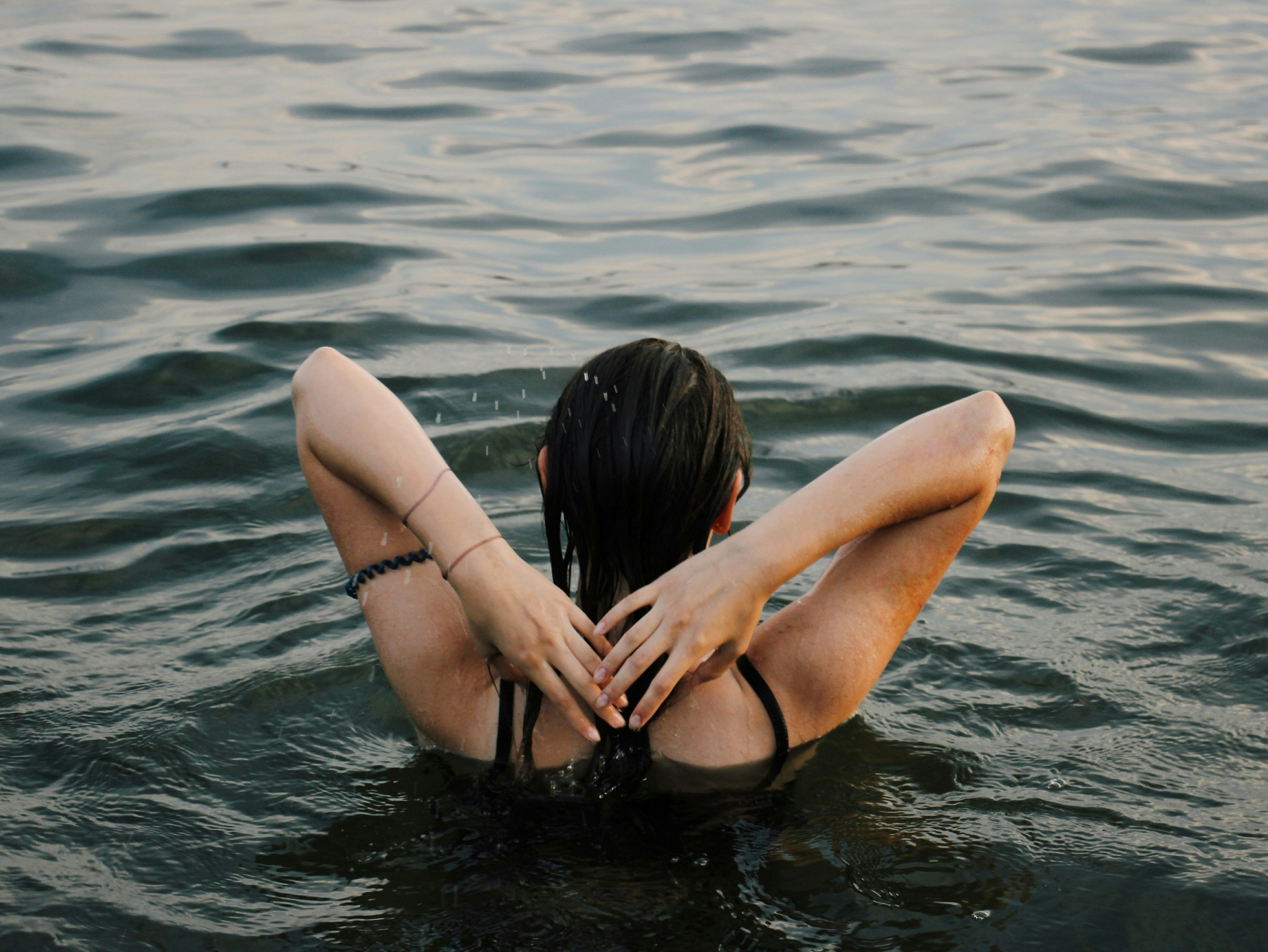 A person with wet hair and tanned skin is partially submerged in water, leaning back with arms extended behind their head, near the water's surface.
