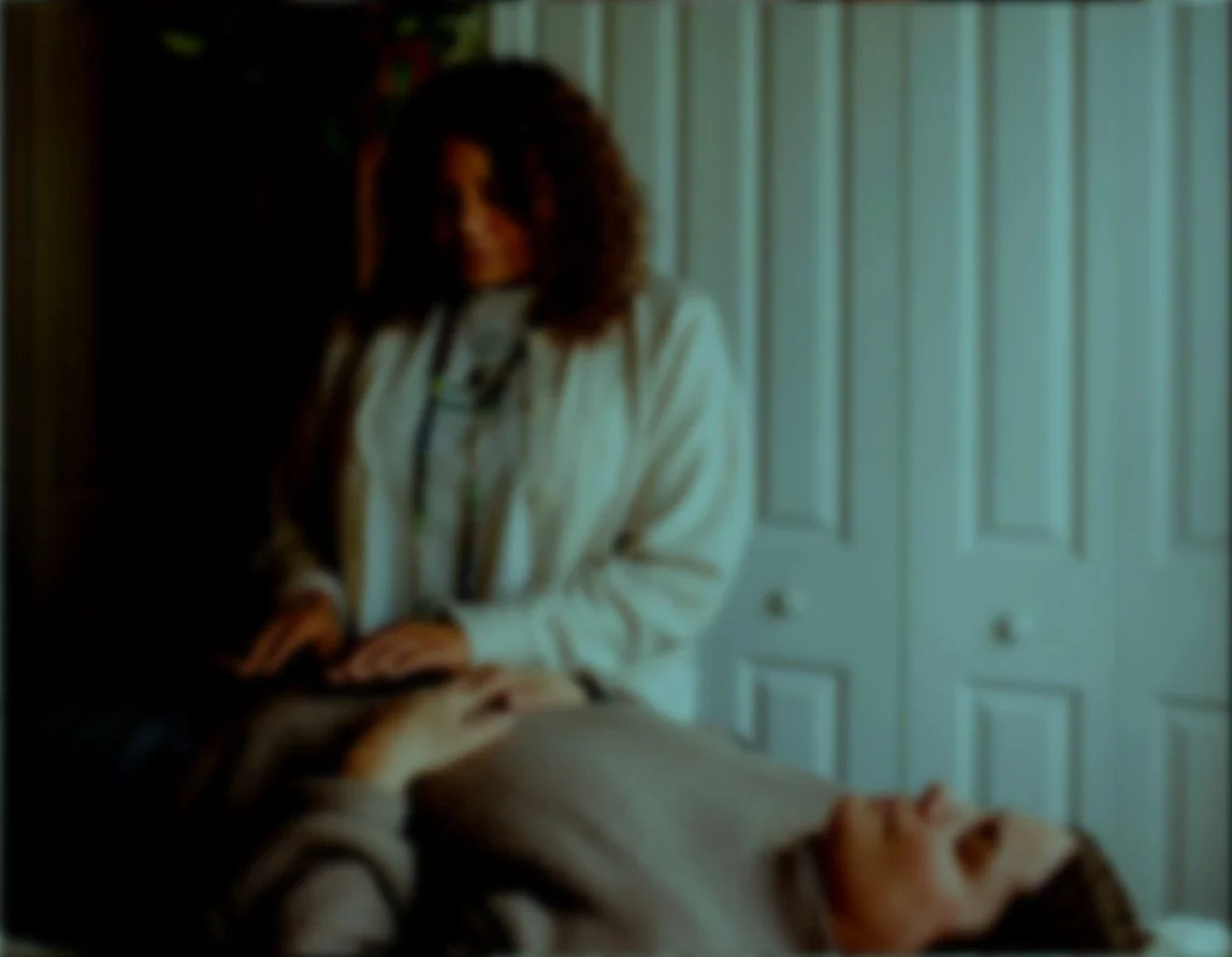 A woman with curly hair standing beside a person lying in a bed, in a room with a closed closet with white doors.