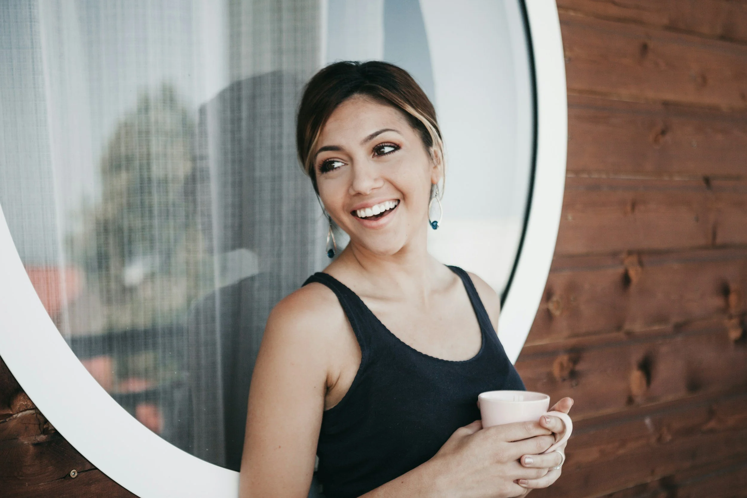 A woman with short brown hair and earrings, wearing a black tank top, smiling and holding a cup while standing next to a round window with a wooden exterior wall.