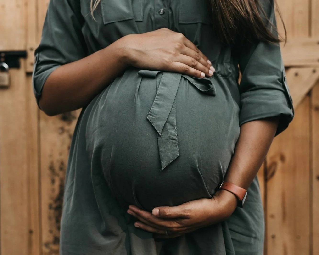 A pregnant woman in a green outfit cradles her baby bump with one hand on top and the other underneath, standing in front of a wooden background.