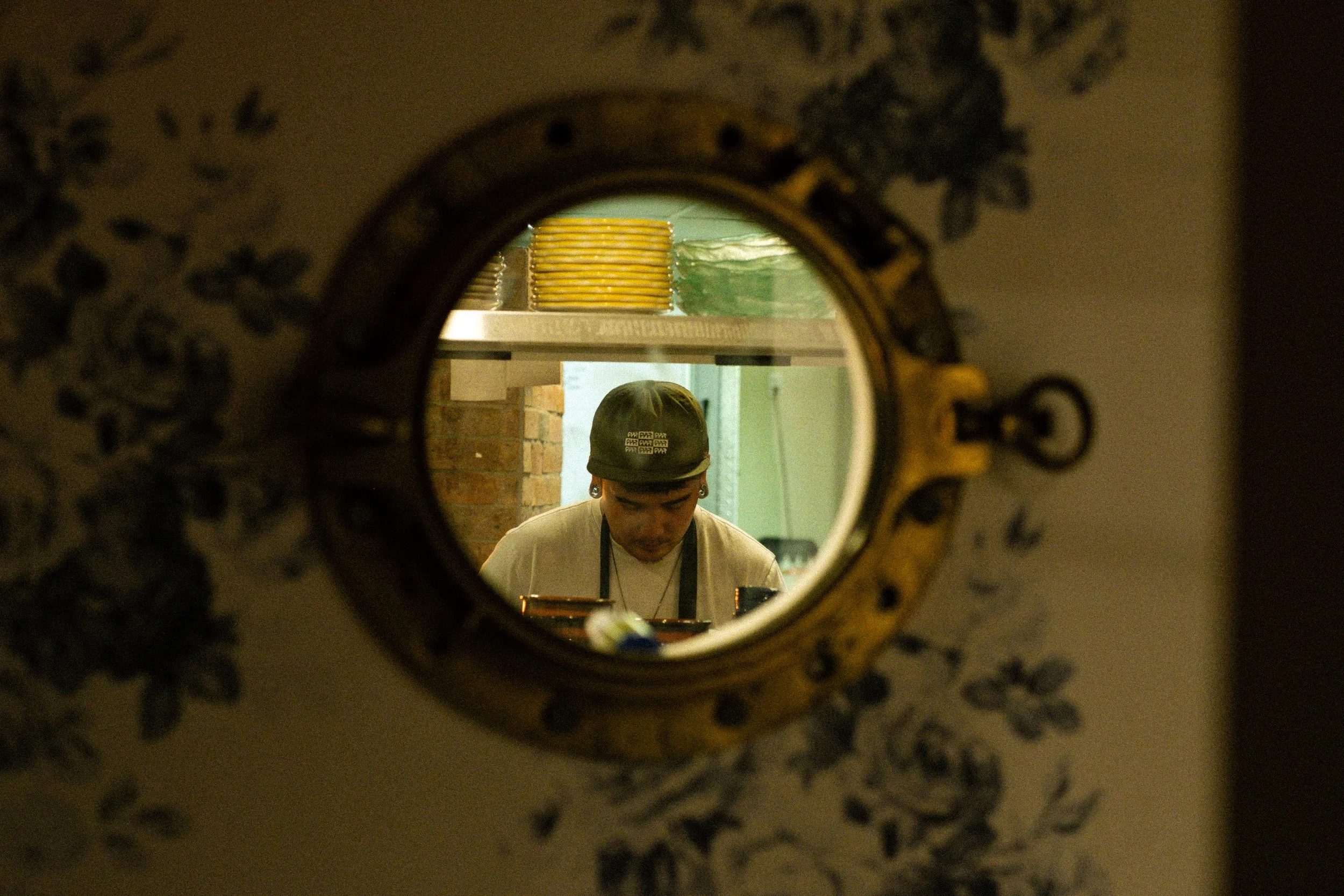 A person wearing a cap and apron seen through a circular mirror, working in a kitchen.
