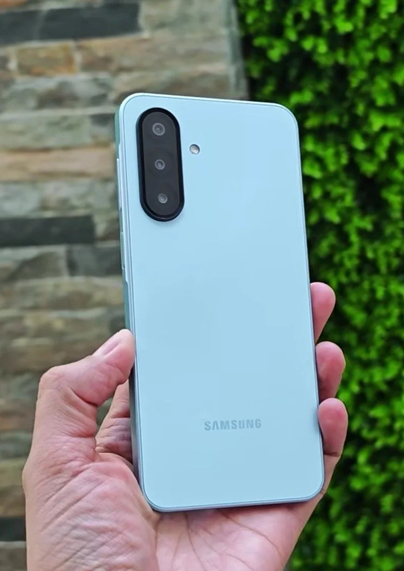 Teléfono inteligente Samsung de color azul en mano, con cámara triple en la parte trasera, fondo de muro de piedra y vegetación.