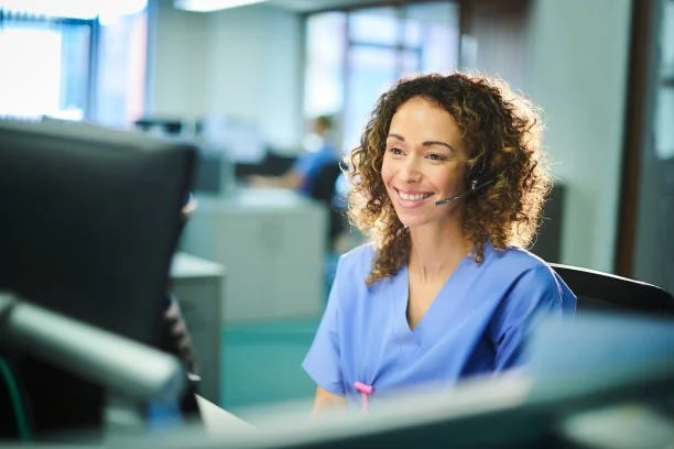 A woman in blue medical scrubs wearing a headset, smiling while working at a computer in a healthcare setting.