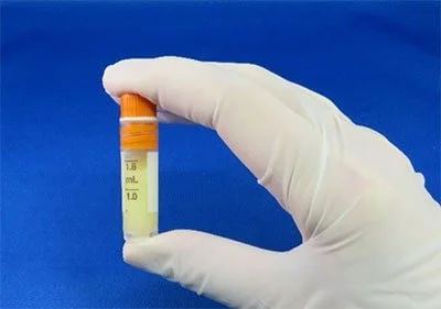 A gloved hand holding a small test tube filled with a yellow liquid against a blue background.