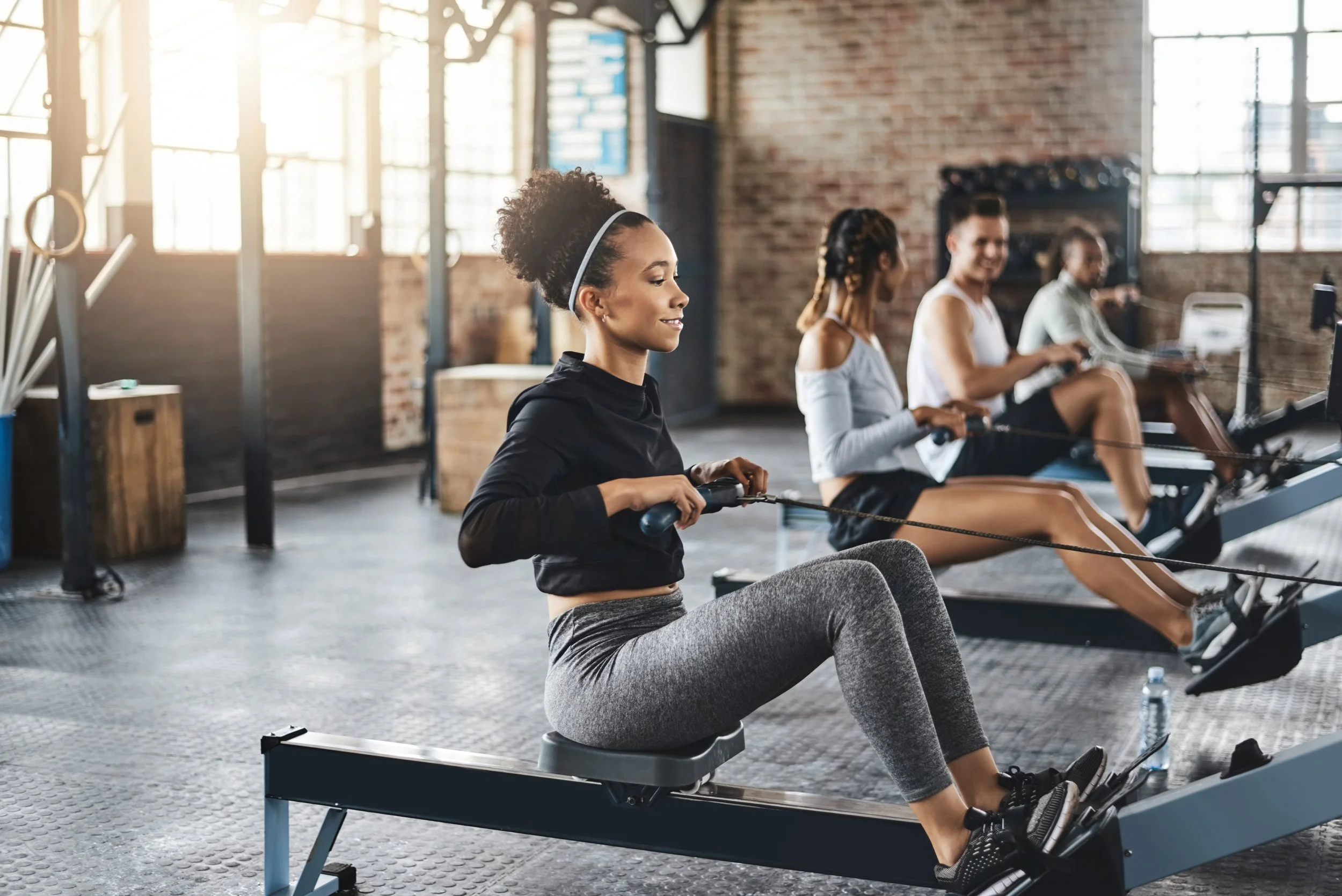 Four women exercising on rowing machines in an industrial-style gym with large windows and brick walls.