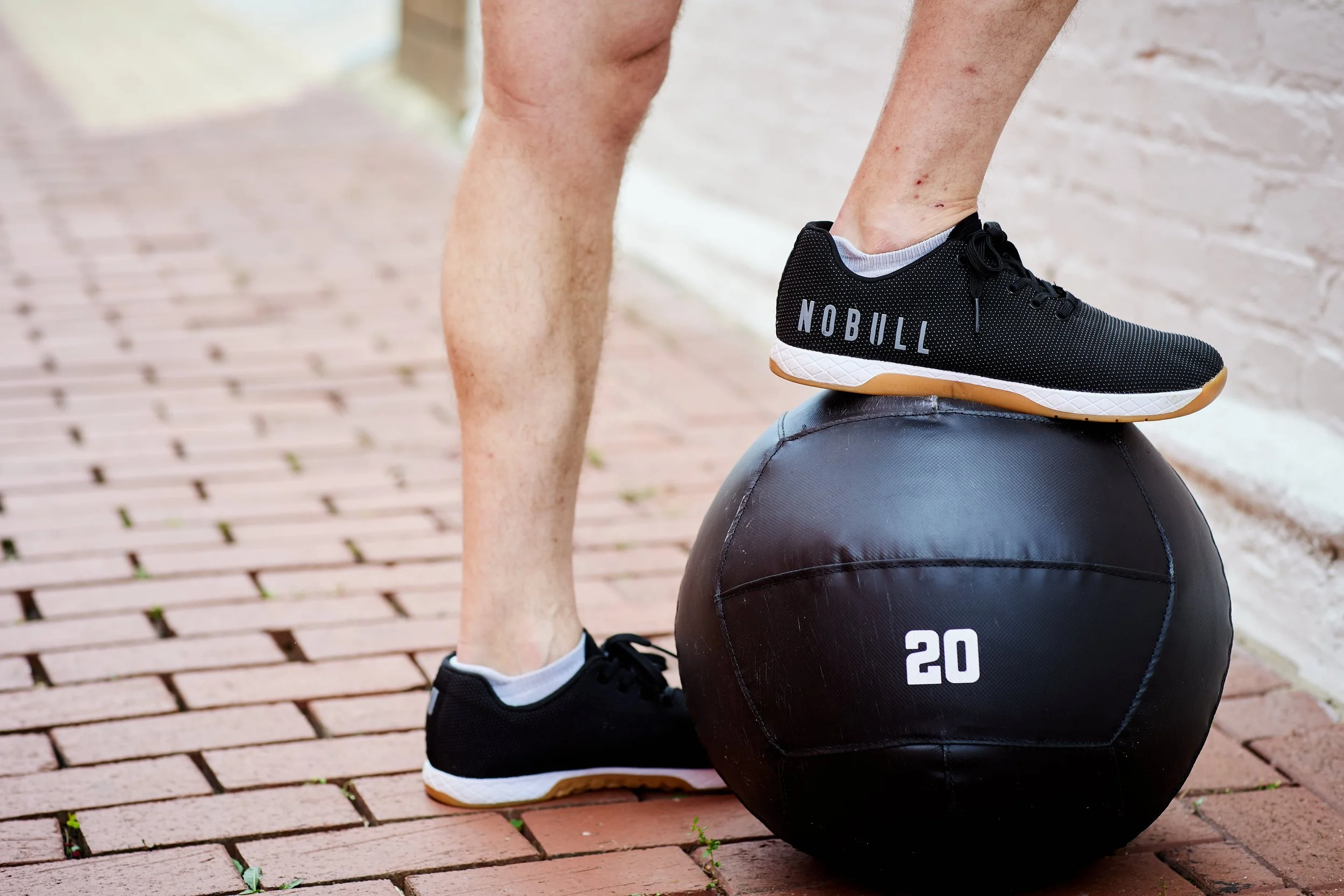 Person standing on one leg with one foot on a black medicine ball with the number 20, outdoors on a brick sidewalk, wearing black athletic shoes with the word NOBULL.