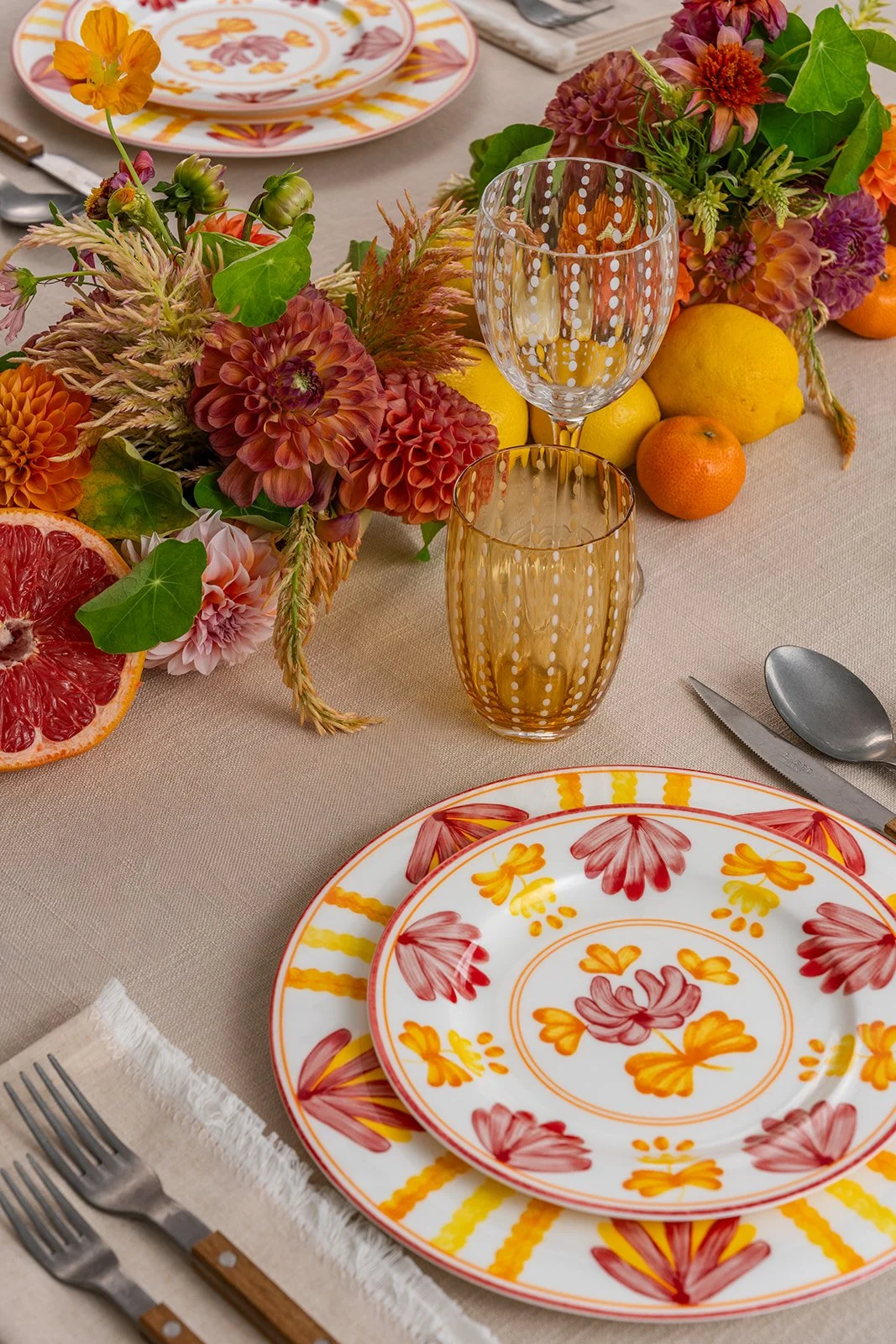 A table setting with floral patterned plates, amber-tinted glassware, and a centerpiece of vibrant flowers, lemons, and citrus fruits on a beige tablecloth.