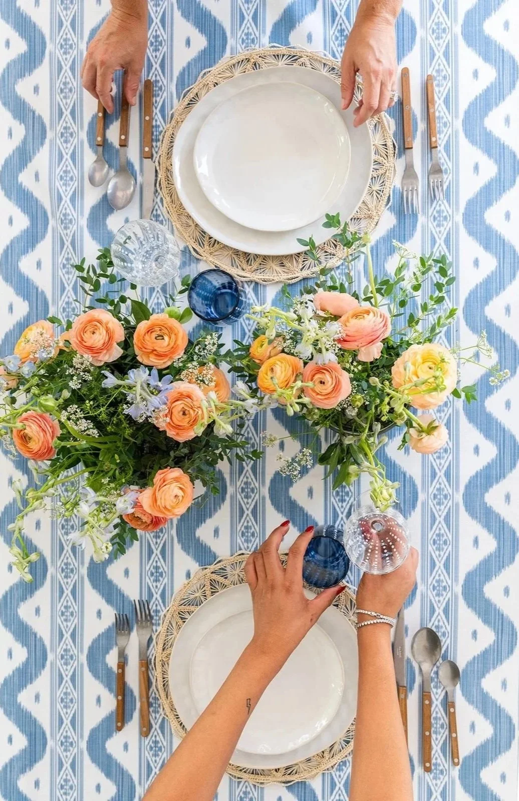 Top-down view of a table setting with two place settings, floral centerpiece, and glasses on a blue and white patterned tablecloth.