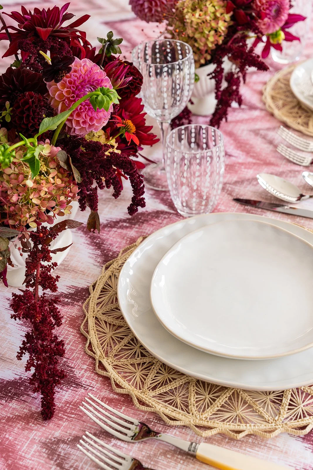 A formal dining table with pink and burgundy floral arrangements, white plates, silver cutlery, and pink textured tablecloth.