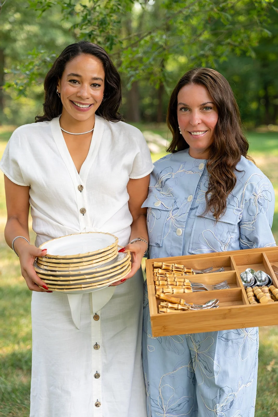 Two women standing outdoors holding serving dishes and utensils, with trees and greenery in the background.