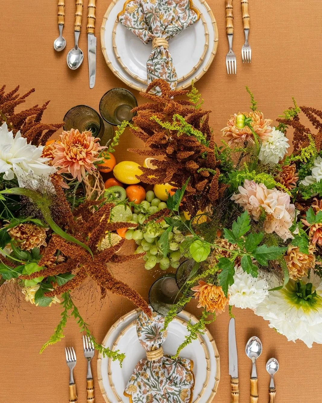 A table setting with floral centerpiece, neatly arranged plates with a patterned napkin, cutlery, and glasses, surrounded by fruits and flowers on a table with a warm orange surface.