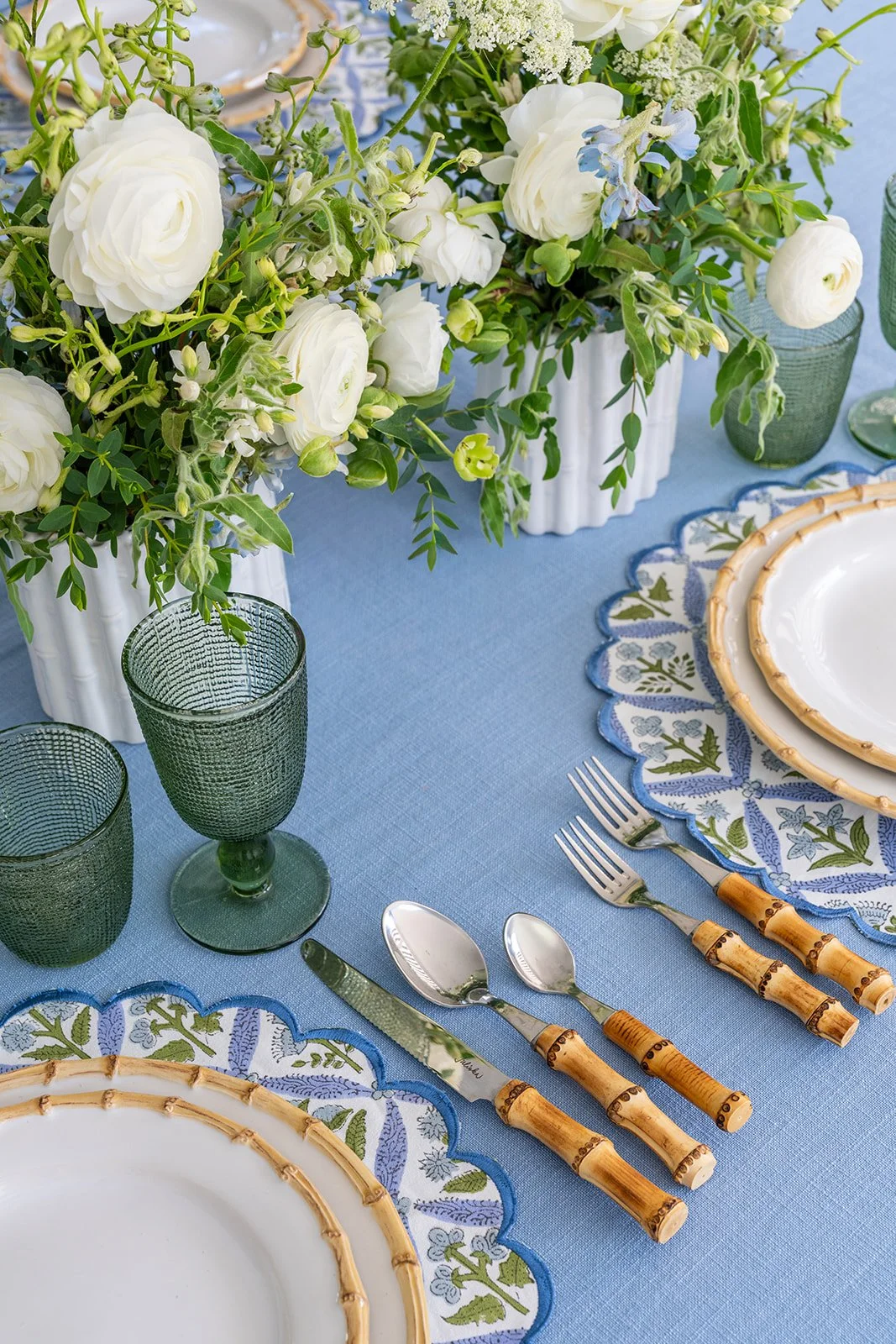 Table setting with white plates, bamboo-handled utensils, green glassware, and floral centerpieces in white vases on a blue tablecloth.