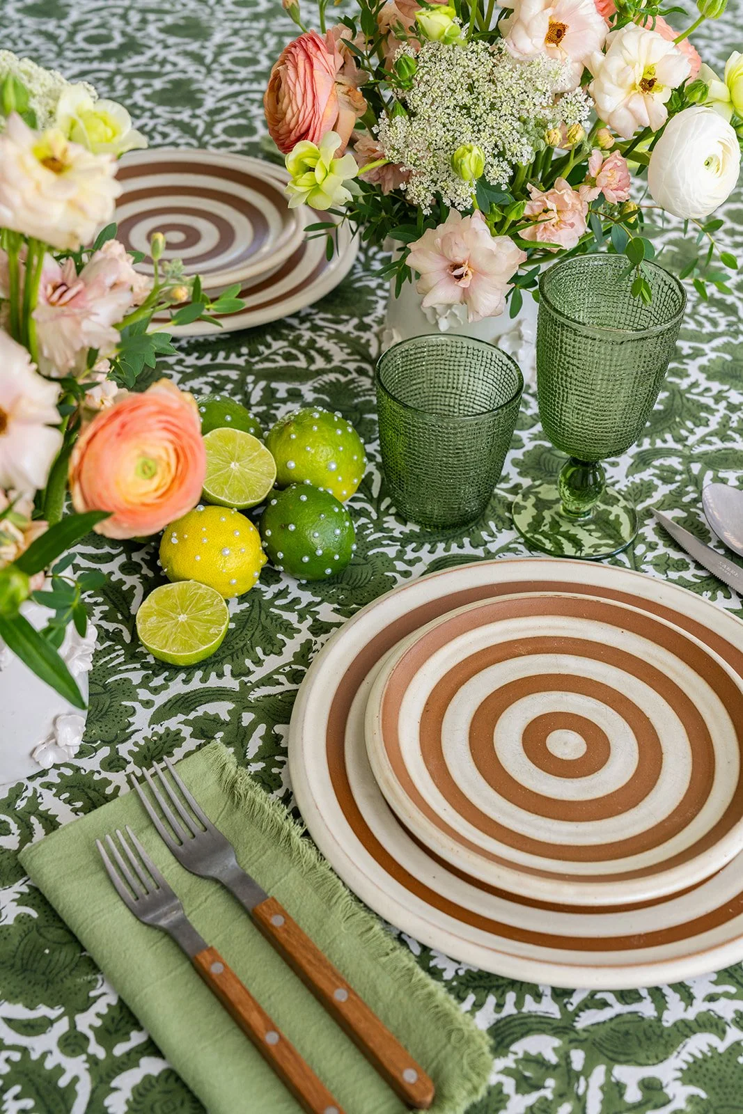 A table setting with floral centerpiece, green glasses, striped plates, cutlery on a green napkin, and decorative limes.
