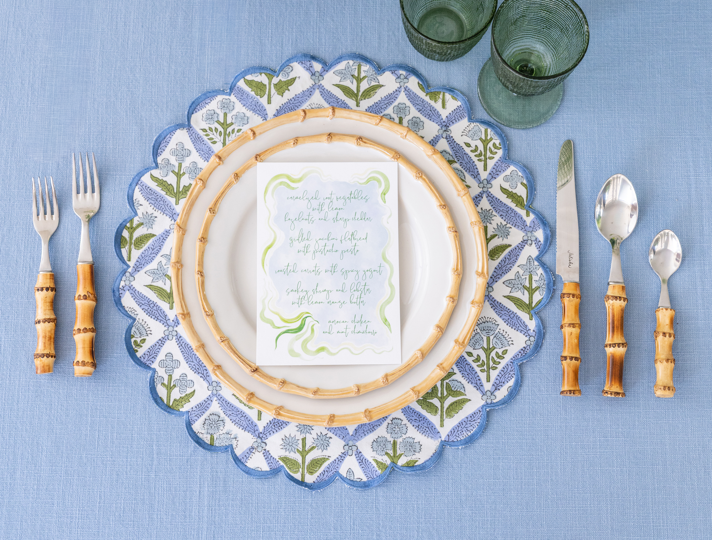 Table setting with two green glasses, a white plate with a woven bamboo rim, a blue and white embroidered placemat, a handwritten menu, a knife, two forks, and three spoons with bamboo handles, on a light blue tablecloth.