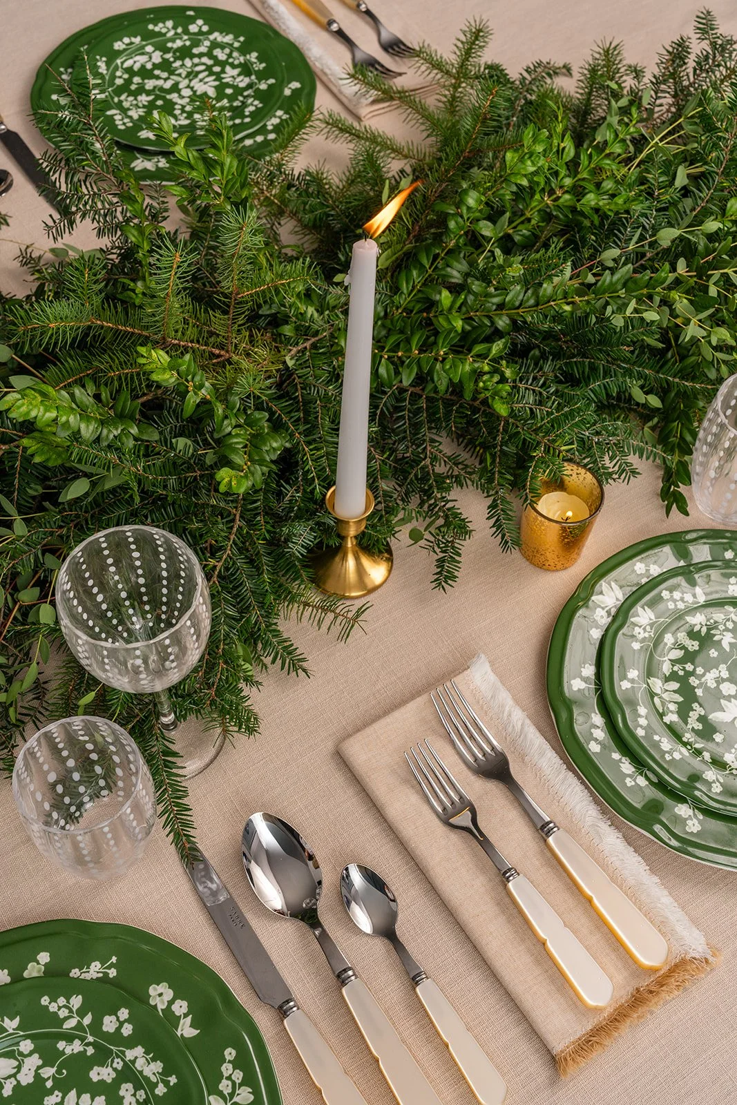 Festive dinner table decorated with greenery, a white candle in a gold holder, gold and white candle in a gold holder, green plates with white floral patterns, glassware with polka dot design, and silverware on a beige napkin.
