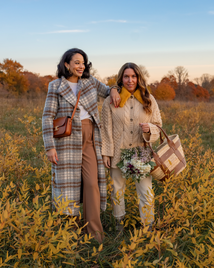 Two women standing in a field of yellow plants during fall, smiling at the camera. One woman has curly hair, wearing a plaid coat and beige pants, with her arm around the other woman. The second woman has long brown hair, wearing a beige knit sweater with a yellow collar, holding a bouquet of flowers and a woven bag.