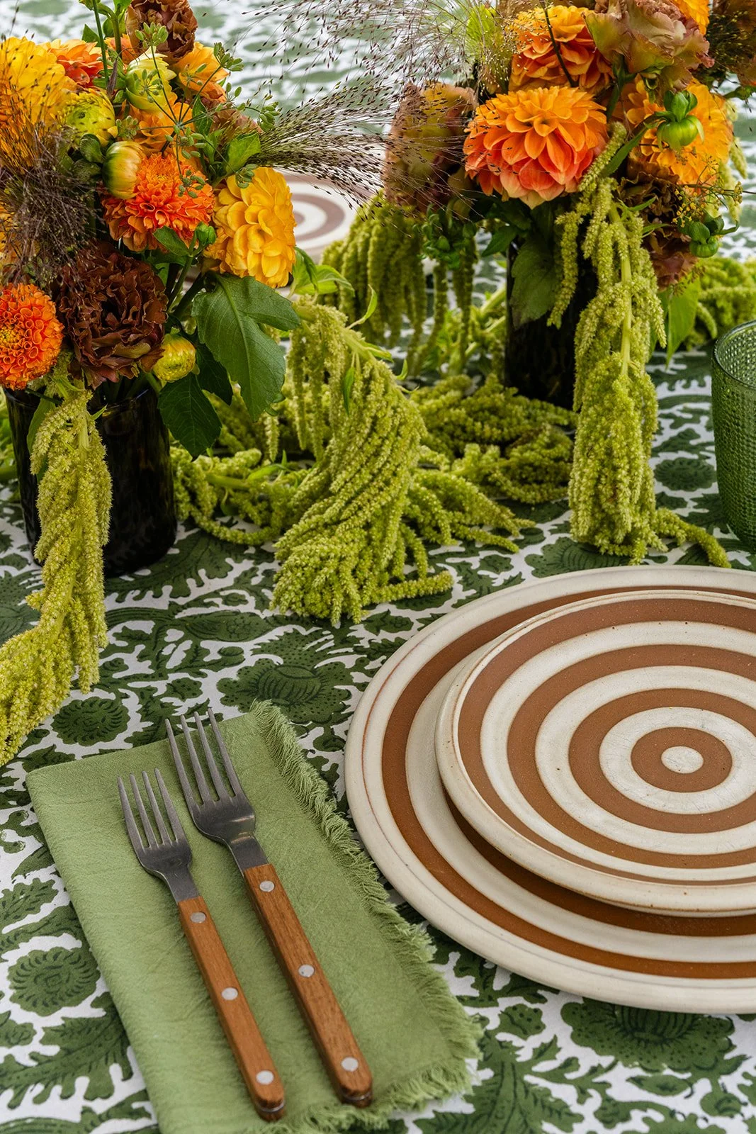 A table setting with floral centerpieces in black vases, featuring orange and yellow flowers, green foliage, and hanging yellow-green amaranth. The table has a green and white patterned tablecloth, a green napkin with two forks, and a set of three concentric circle patterned brown and white plates.