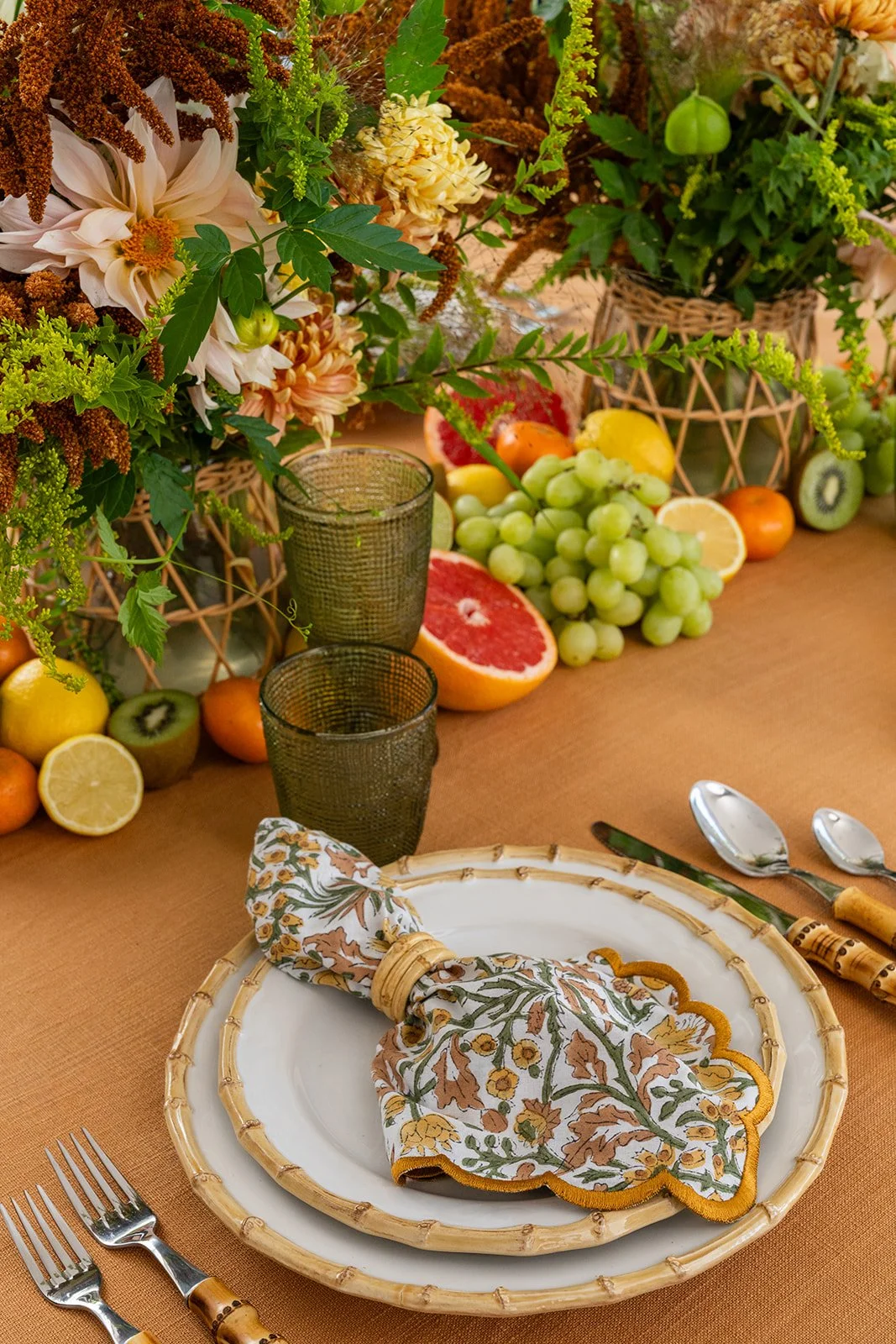 A table set with a floral arrangement, grapes, citrus, and other fruits, with a white plate, napkin, and cutlery.