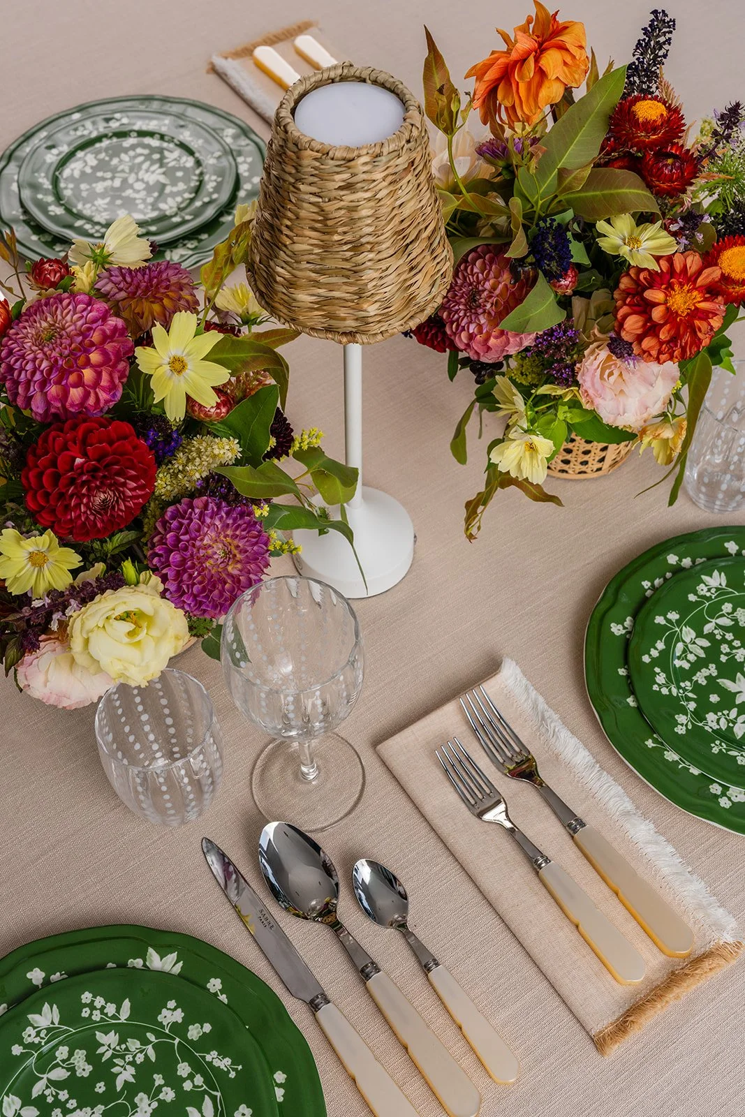 Table setting with green floral plates, silverware with cream-colored handles, clear patterned glasses, floral centerpieces with pink, red, orange, yellow, and purple flowers, a wicker lamp with a white stand, a beige tablecloth, and a napkin with fringed edges.