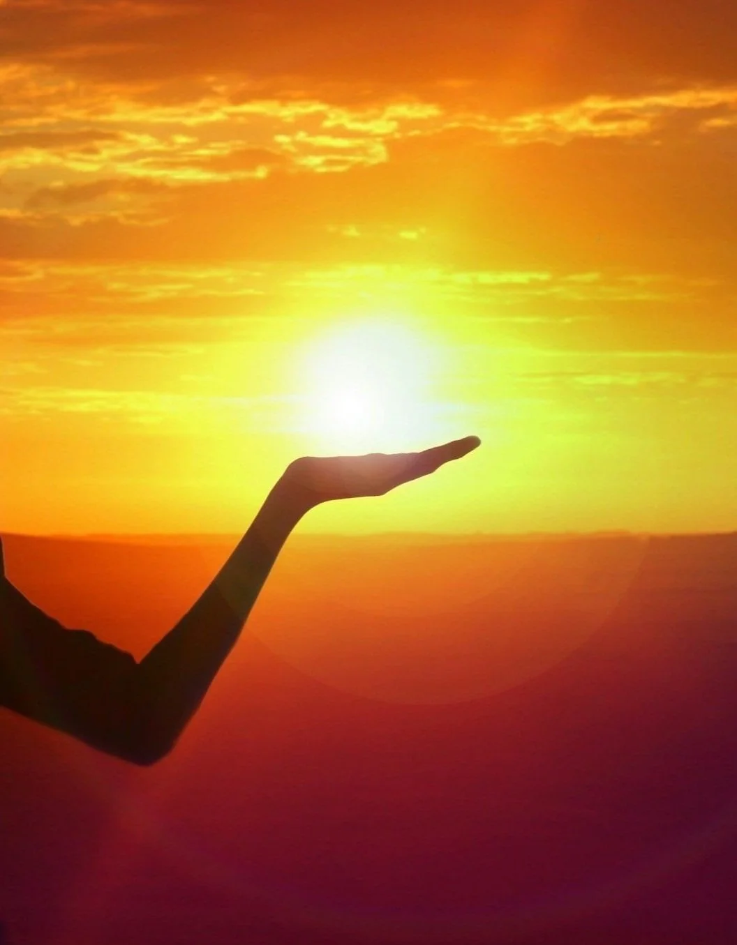 A person's silhouette appears to hold the sun in their hand during a bright, colorful sunset with orange and yellow clouds.