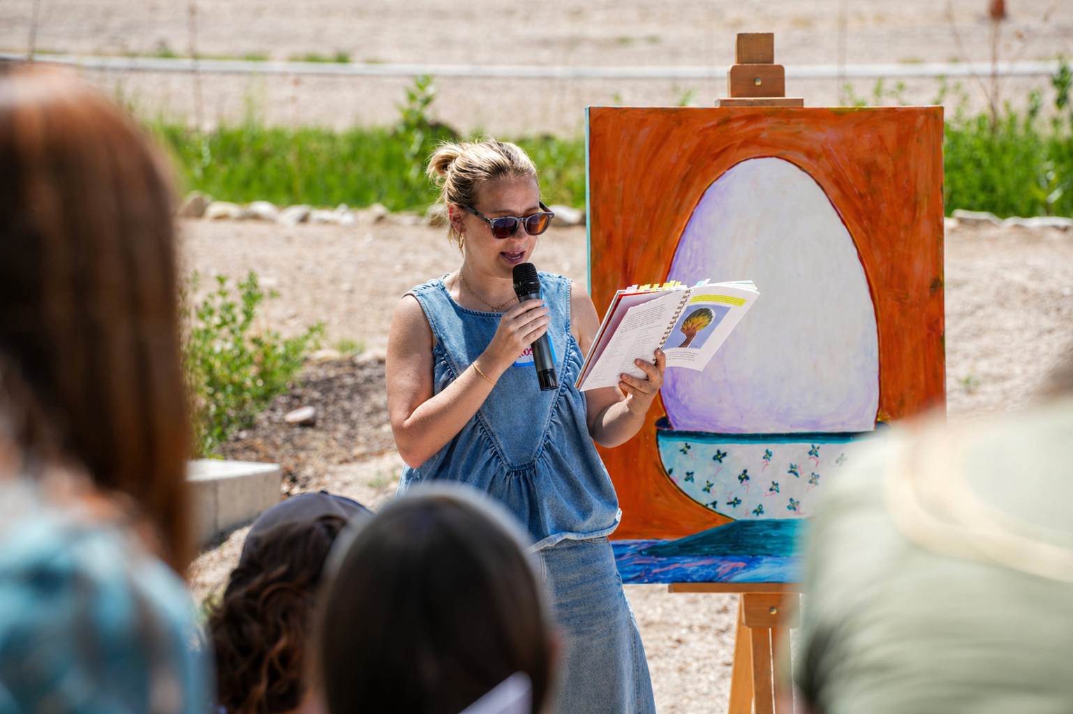 Woman reading aloud from a book during an outdoor event, standing in front of an abstract painting on an easel, with an audience in the foreground.
