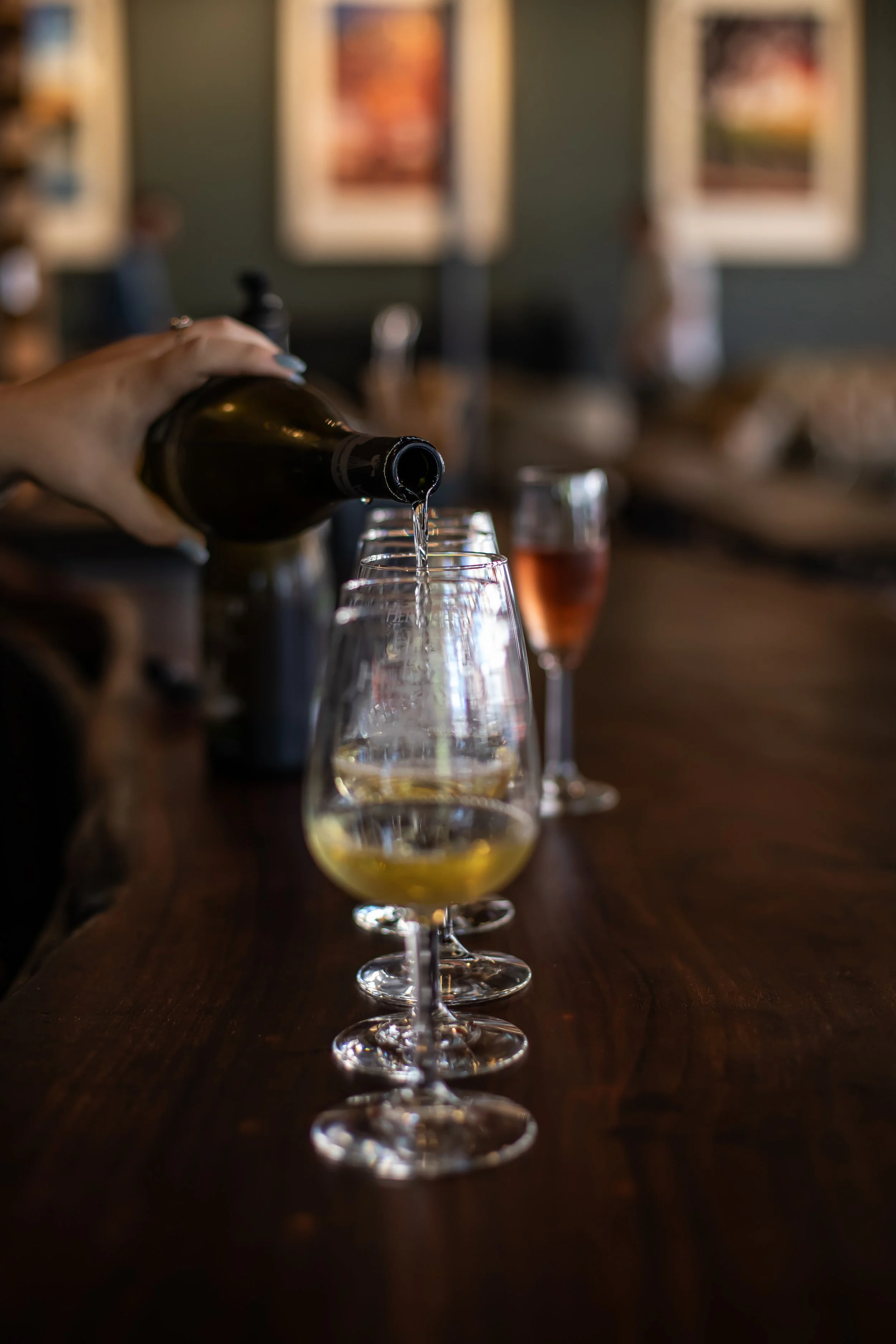 A person pouring a glass of white wine at a bar, with additional glasses and a glass of rosé in the background.