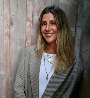 Dr. Tal Ginsburg, smiling, with shoulder-length blonde hair, wearing a checkered blazer and layered necklaces, standing against a wooden wall.