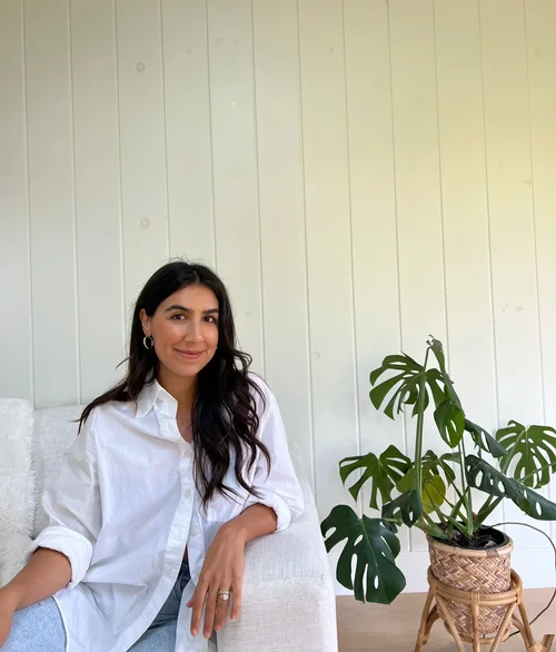 Dr. Chelsea Sarai, founder of Brentwood Therapy Collective, sitting on a white couch with a white wall background, wearing a white shirt, next to a large potted plant with green leaves.
