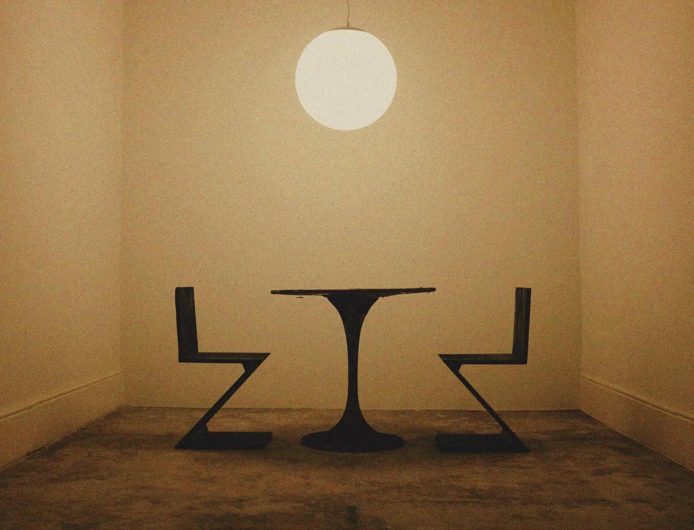 A small round table with two chairs placed on either side, with the chairs formed by the shape of the letters 'Z' and 'S', in a room with beige walls and a round ceiling light.
