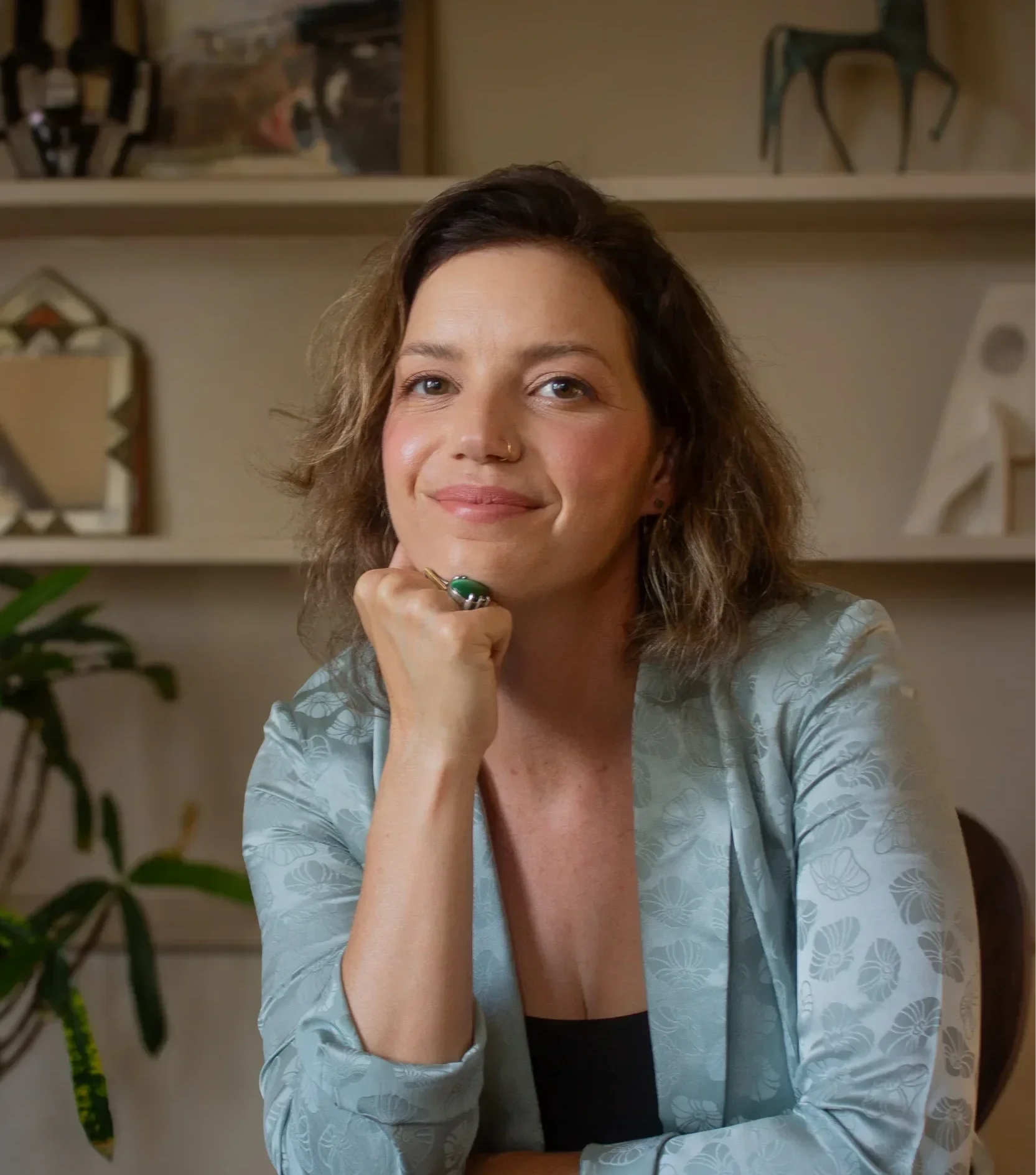Kim Brounstein, LMFT, wearing a light blue patterned blazer and black top, sitting at a table in a cozy home interior with shelves and artwork in the background, smiling softly at the camera.