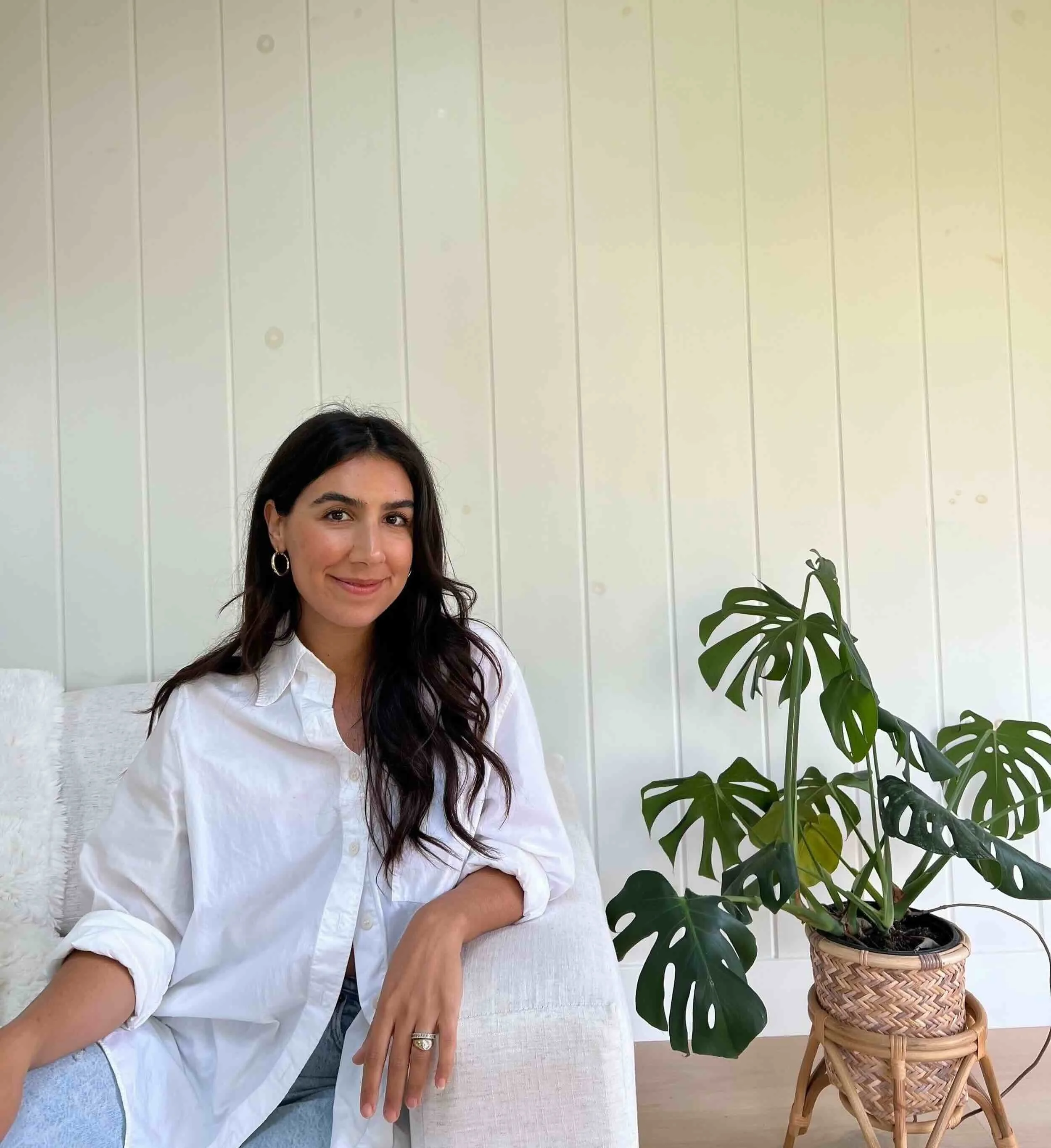 Dr. Chelsea Sarai, founder of Brentwood Therapy Collective, sitting on a white couch with a white wall background, wearing a white shirt, next to a large potted plant with green leaves.