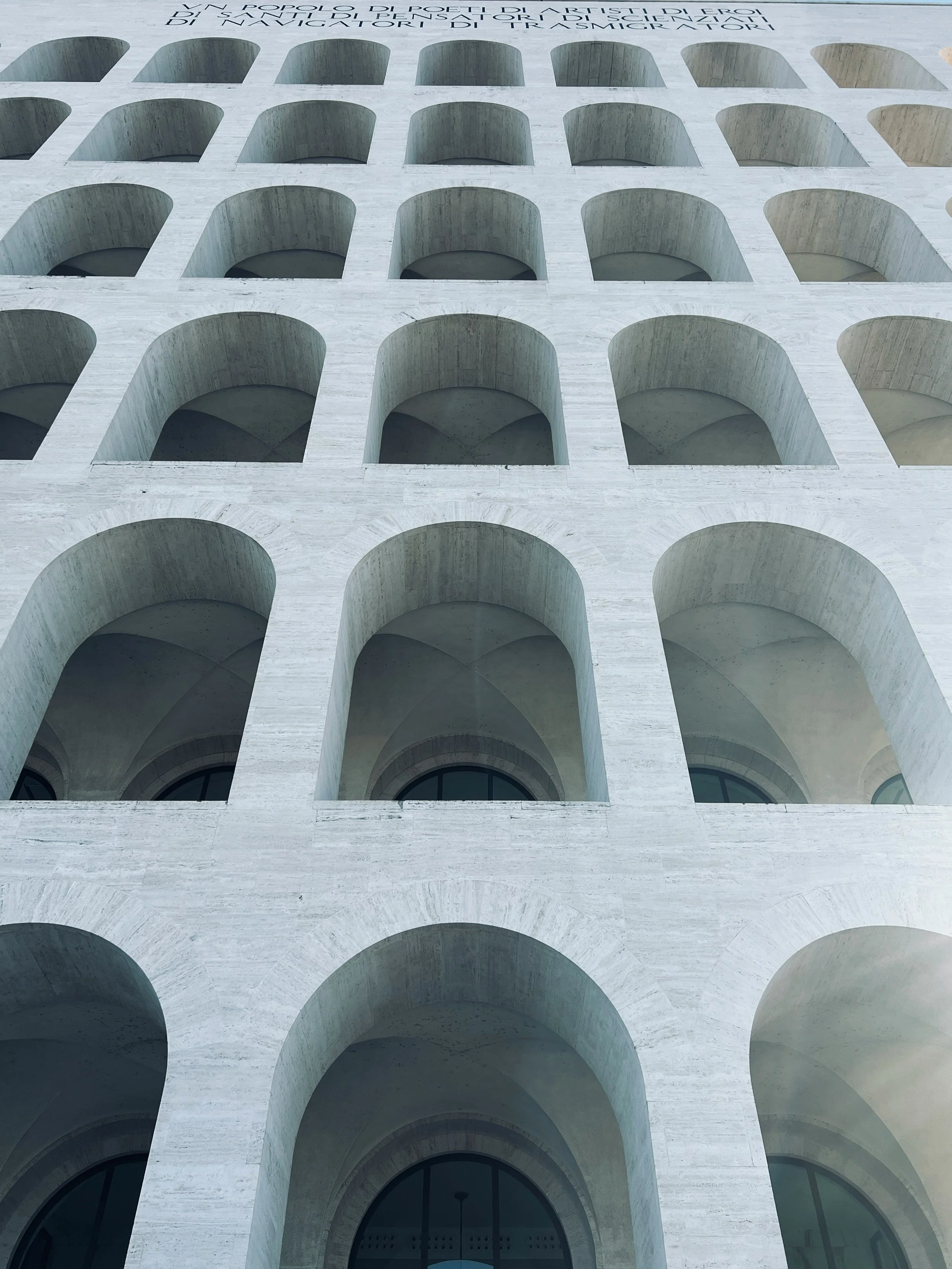 A modern building facade with multiple large, arched openings arranged in a grid pattern. The building appears to be made of light-colored stone or concrete, and the image is taken from below, looking upward.