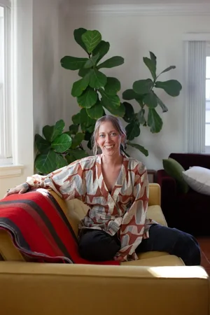 Dana Kurth, smiling at the camera, on a yellow couch with a large indoor fig tree behind her.