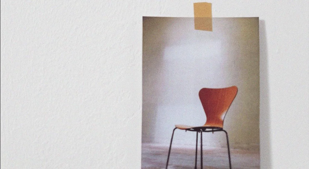 Empty wooden chair with black metal legs in a small nook with white walls and a piece of yellow tape at the top.