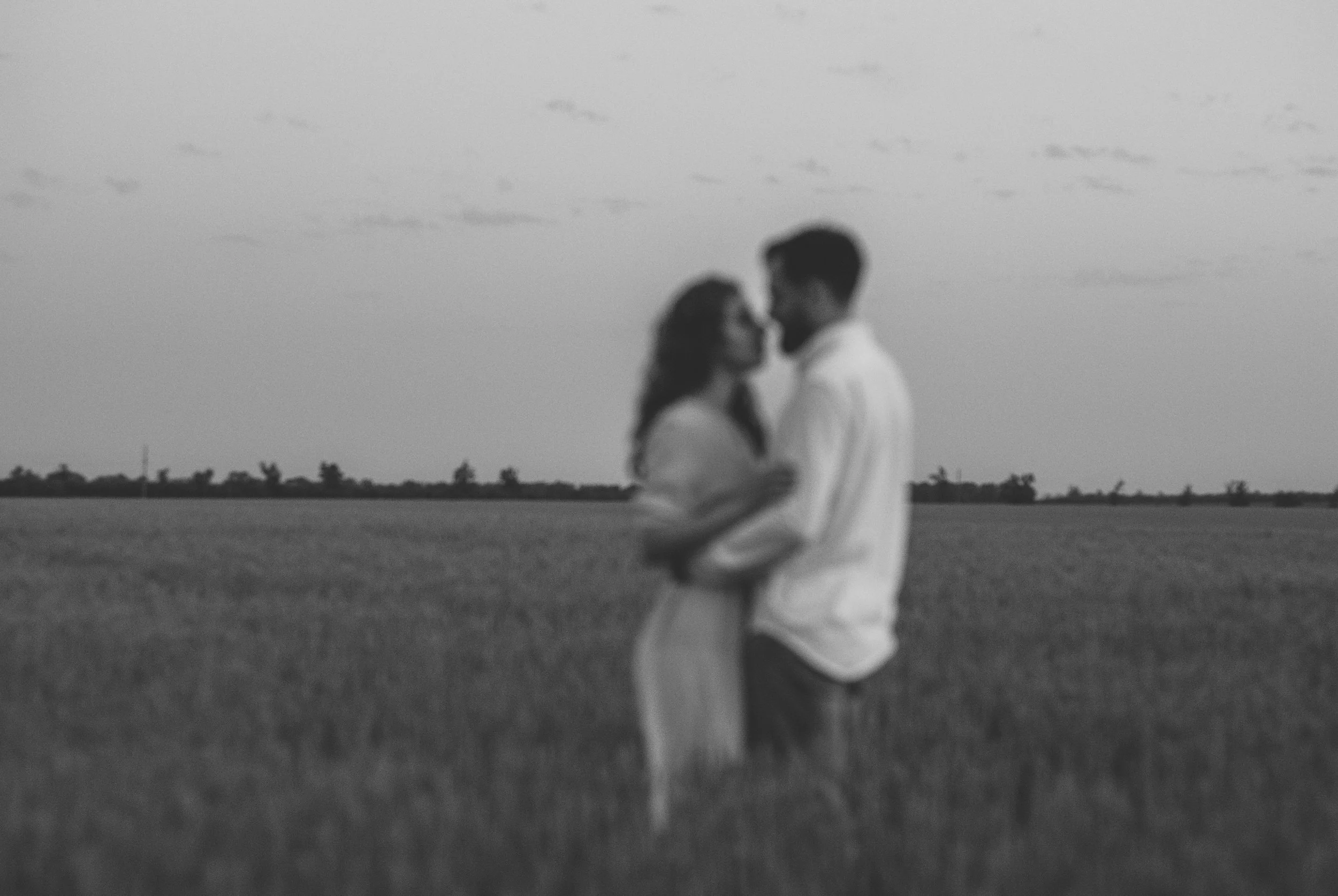 Black and white photo of a couple standing close to each other outdoors with a distant tree line, holding hands, with their faces near each other, in a serene landscape.