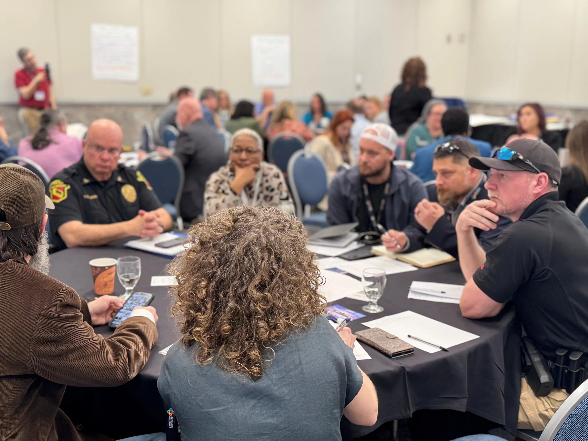 A group of diverse people gathered around tables during a meeting or conference, with some taking notes and listening attentively, while a woman stands at the back addressing the group.