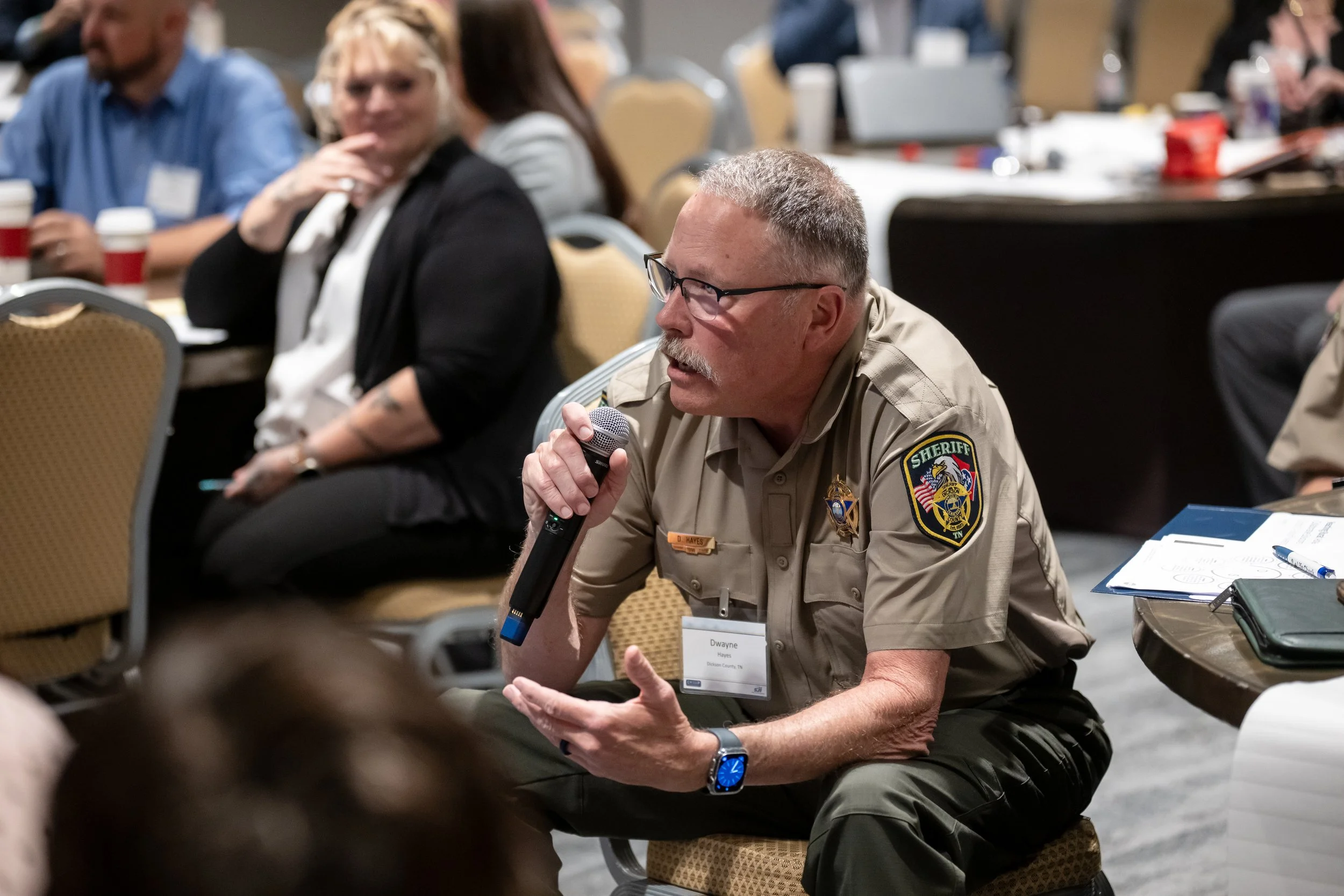 A middle-aged man with glasses and a mustache, wearing a sheriff's uniform, sitting on a chair, holding a microphone, and speaking during a conference or meeting. Several people are sitting behind him, listening and watching.