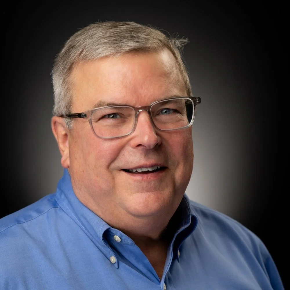 A middle-aged man with glasses, gray hair, wearing a blue collared shirt, smiling in a professional portrait.